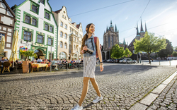 Frau spaziert auf gepflastertem Platz vor historischen Gebäuden bei Sonnenschein.