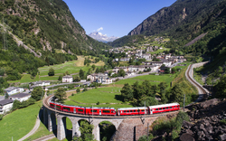 Roter Zug auf einem Viadukt in bergiger Landschaft mit Dorf im Hintergrund.