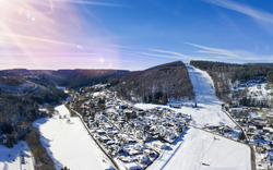Verschneites Dorf in hügeliger Winterlandschaft unter blauem Himmel.
