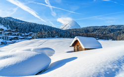 Verschneite Landschaft mit Hütte und Bergen im Hintergrund bei blauem Himmel.